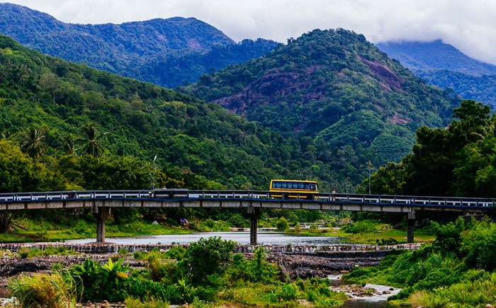 Es una imagen que muestra un autobús amarillo en un puente cruzando un río, con montañas en el fondo, en una carretera rural tailandesa. El paisaje es muy verde y exuberante