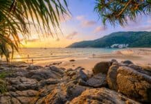 Atardecer en Nai Harn Beach, Phuket, con un cielo naranja, nadadores, personas en la playa arenosa, veleros en el mar y rocas a lo largo de la costa.