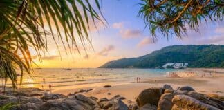 Atardecer en Nai Harn Beach, Phuket, con un cielo naranja, nadadores, personas en la playa arenosa, veleros en el mar y rocas a lo largo de la costa.